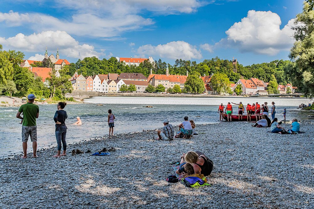 Das Foto zeigt eine Rafting-Gruppe mit orangefarbenen Westen am Kiesstrand des Lechs in Landsberg am Lech, mit der Altstadt (rote Dächer) und grünen Bäumen im sonnigen Hintergrund.