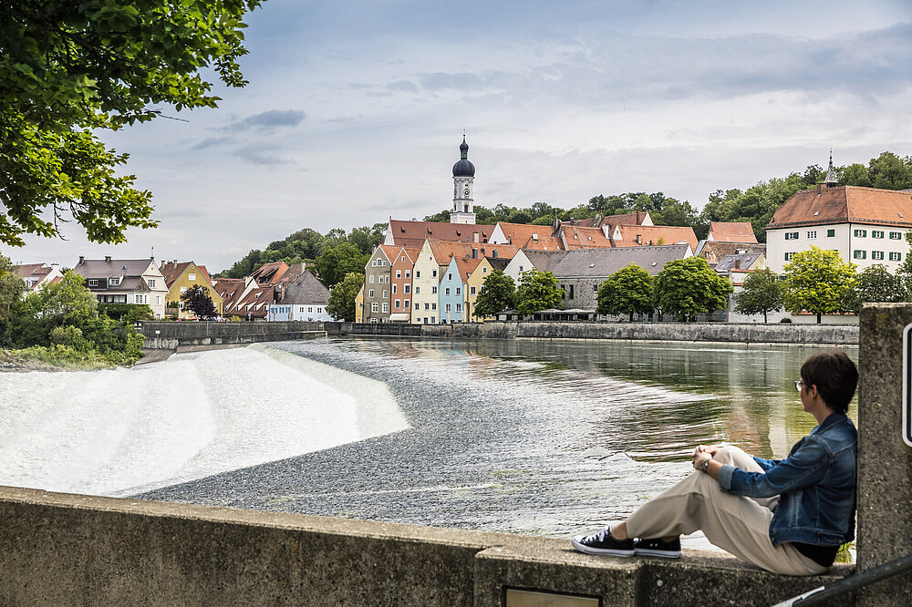 Das Foto zeigt eine junge Frau in Jeansjacke, die auf der Lechwehr-Mauer in Landsberg am Lech sitzt und liest, mit Panoramablick auf die bunte Altstadt, rote Dächer, Kirchturm und Lech-Fluten bei sonnigem Wetter.